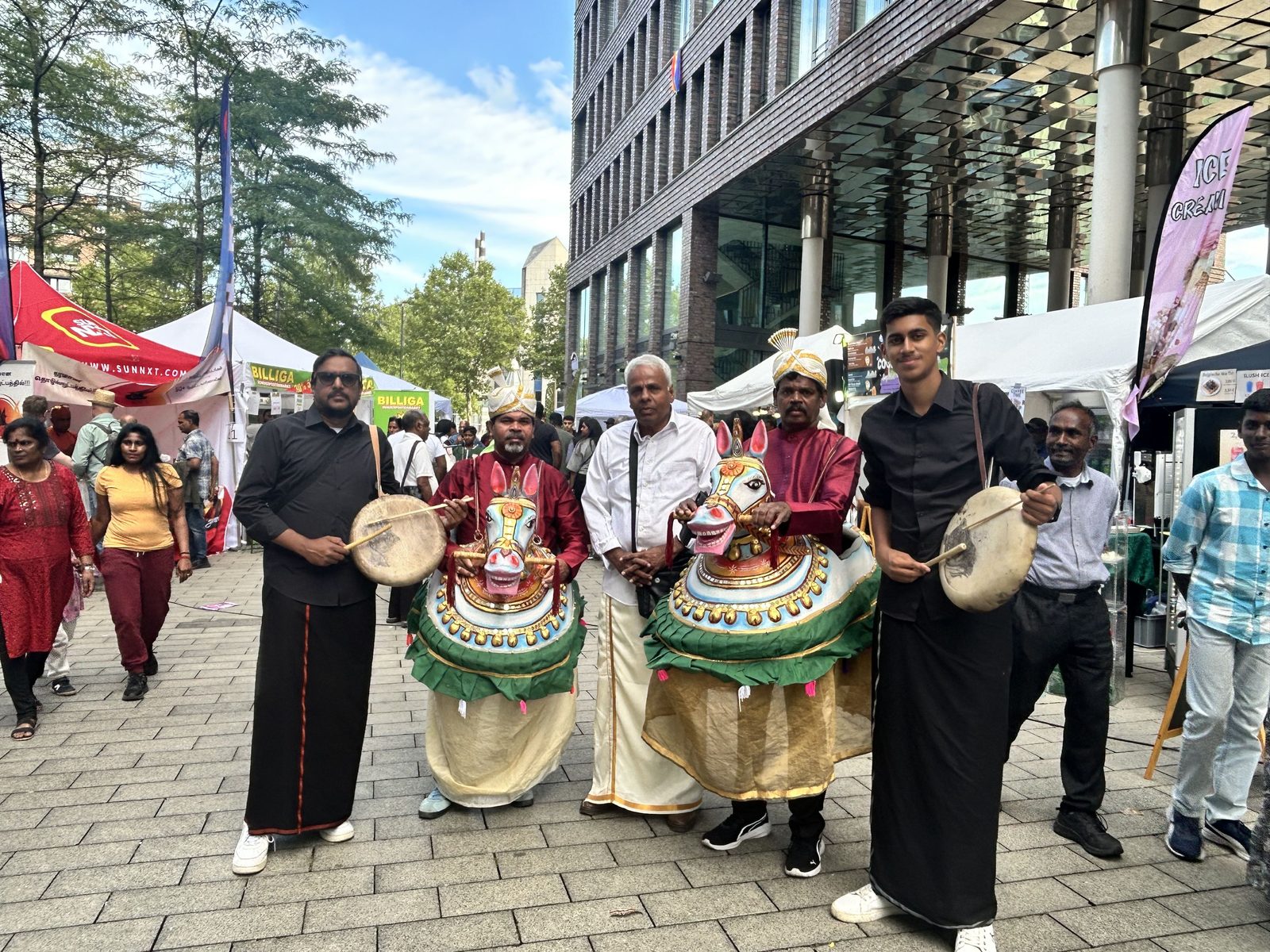 Two Poi Kal Kuthirai dummy horse performers with drummers among the stalls of a street festival in Germany