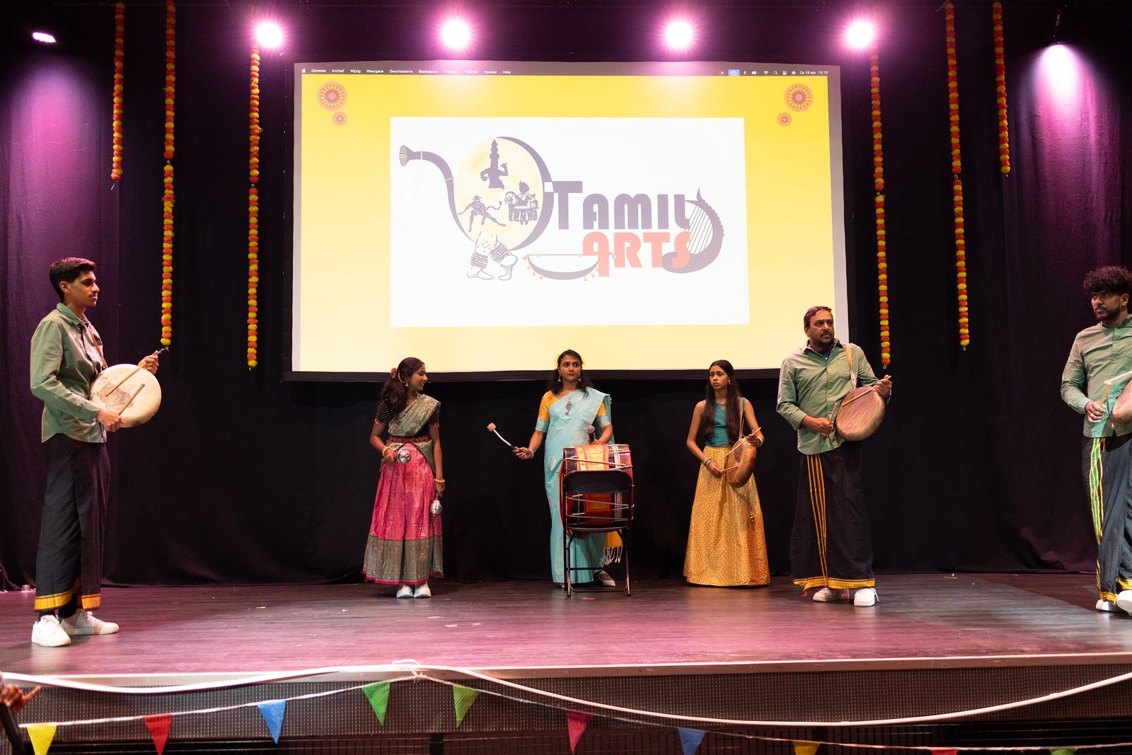 Performers with parai drums on stage in the Netherlands, the Tamil Arts logo projected behind them