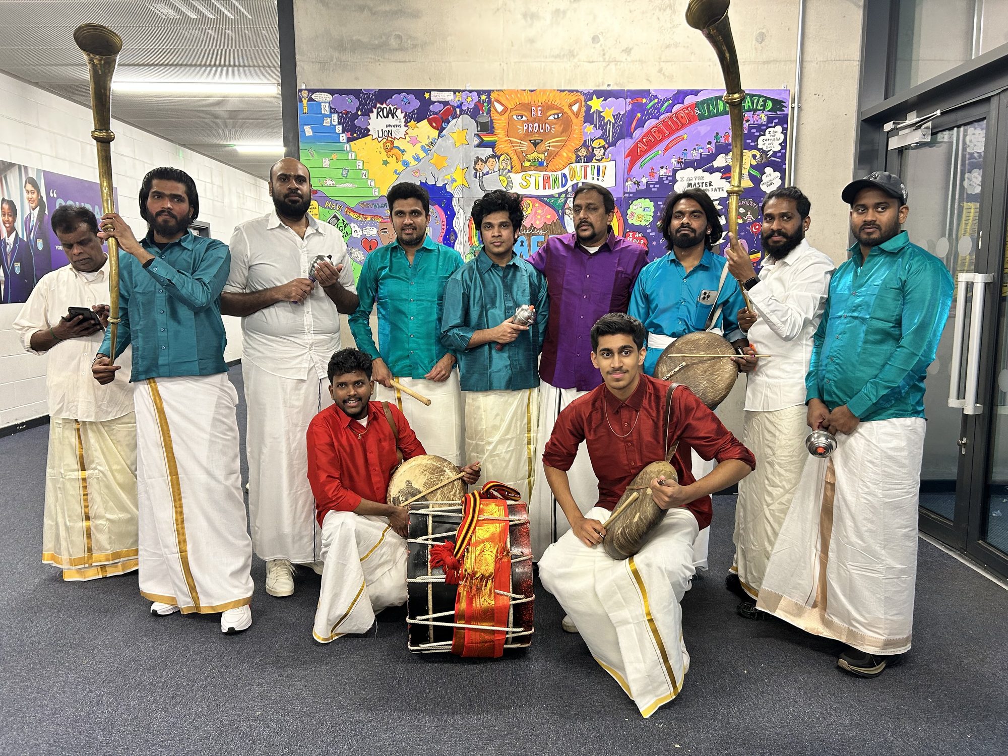 Parai drummers and nadaswaram players posed in a school corridor in front of a student mural