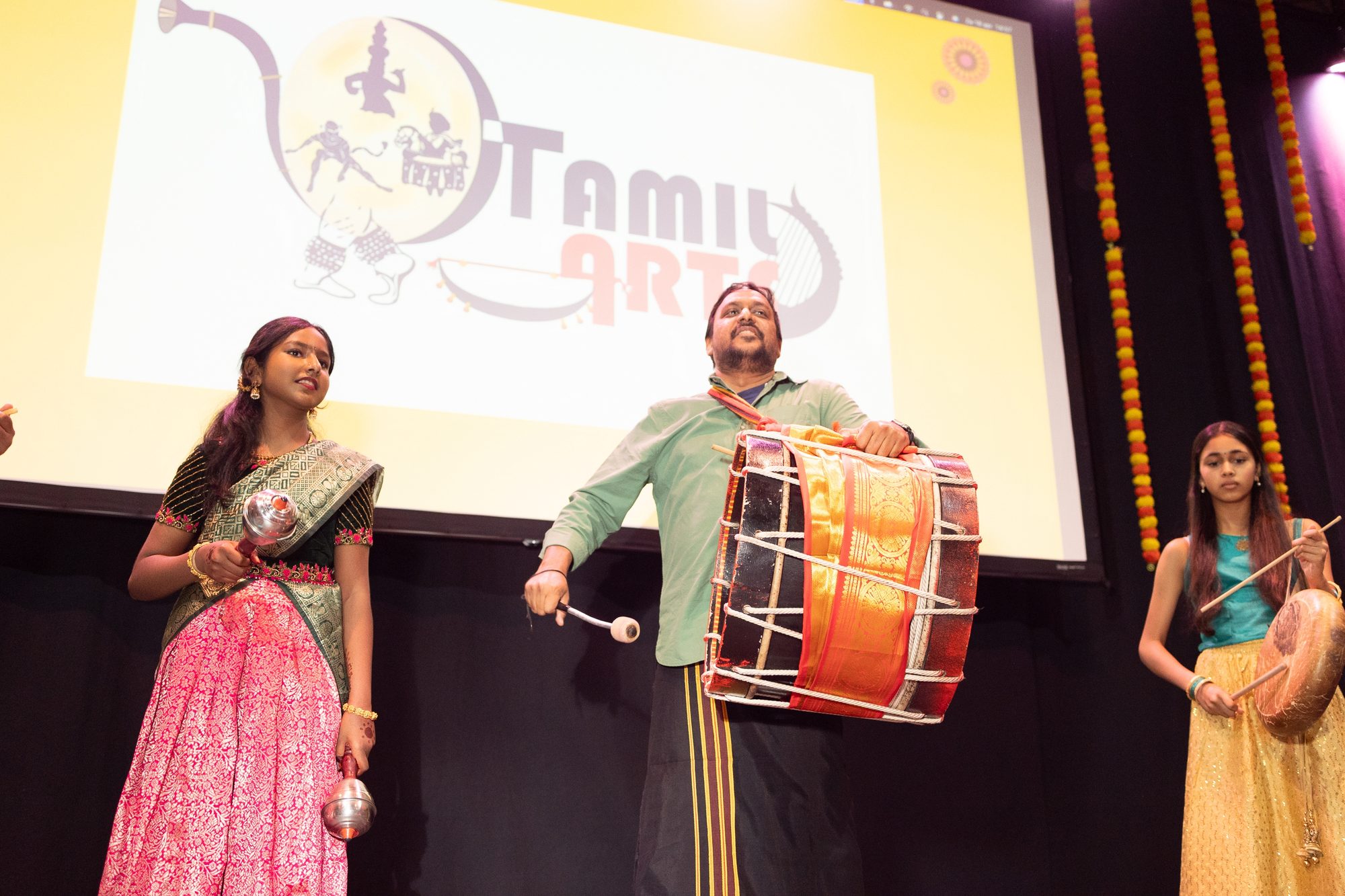A dhol player mid-beat flanked by dancers under the Tamil Arts logo backdrop