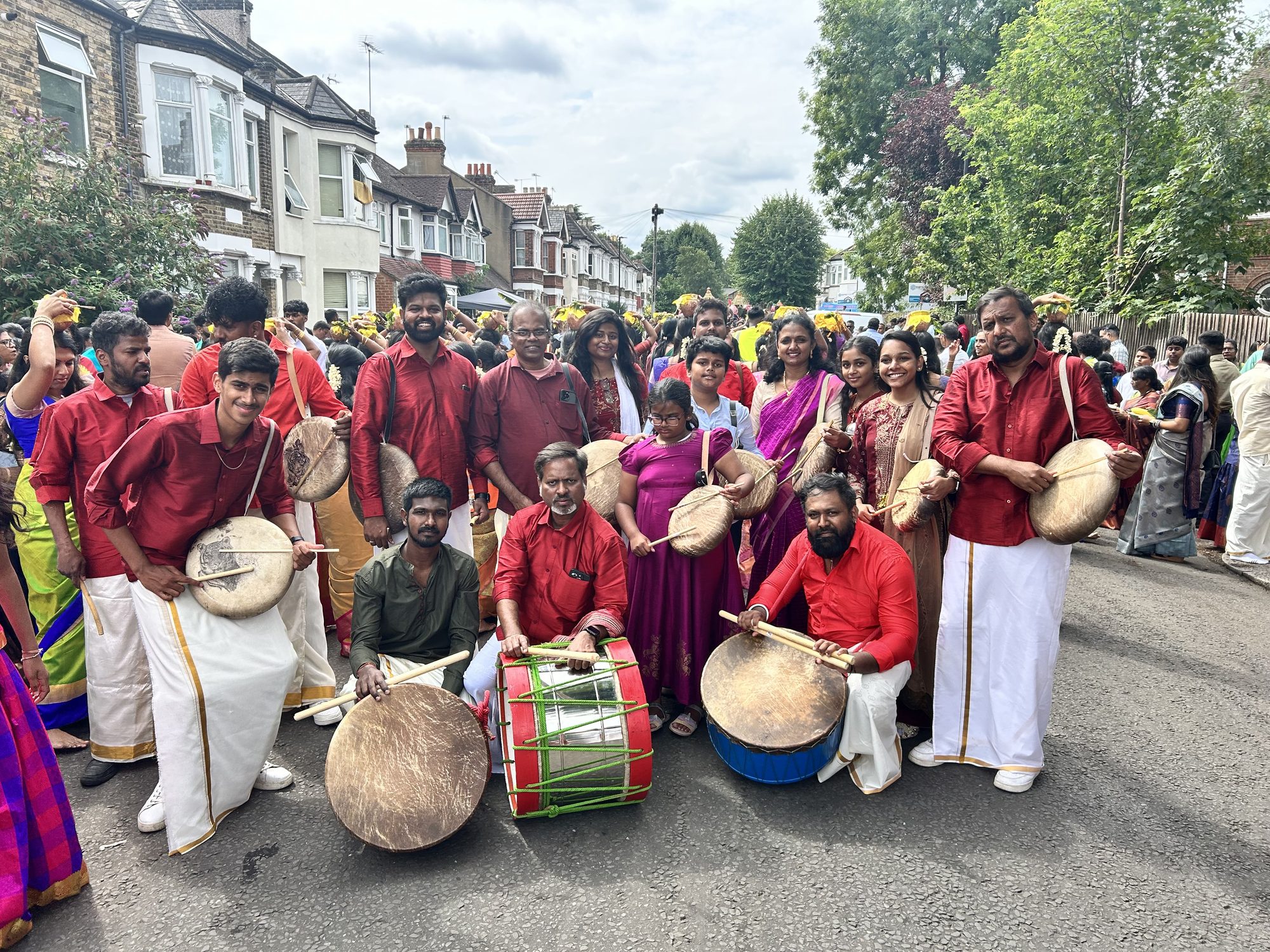 A large group of red-shirted parai drummers with families at a temple procession on a London residential street