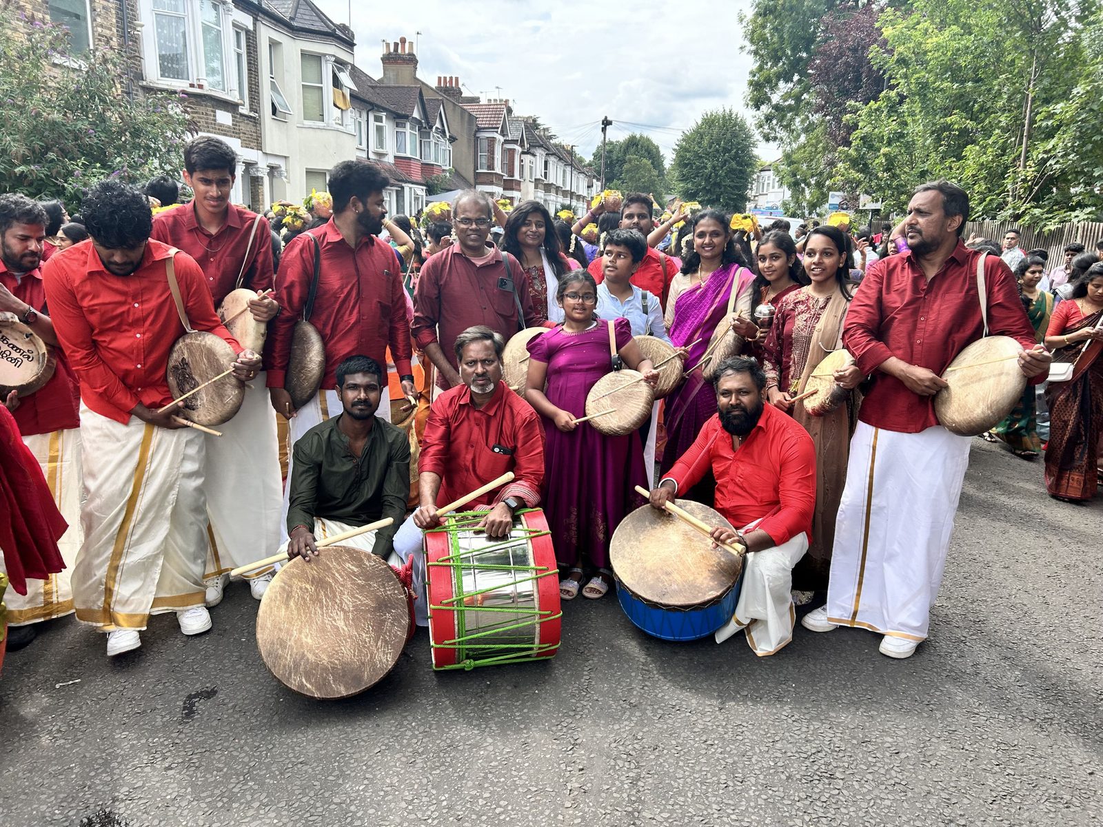 Parai drummers in red seated and standing in the road during a temple procession, kavadi carriers behind them