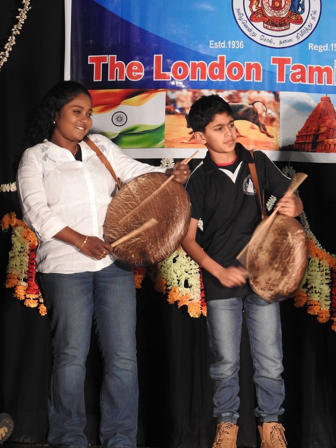 Two young parai drummers performing in front of a London Tamil Sangam banner