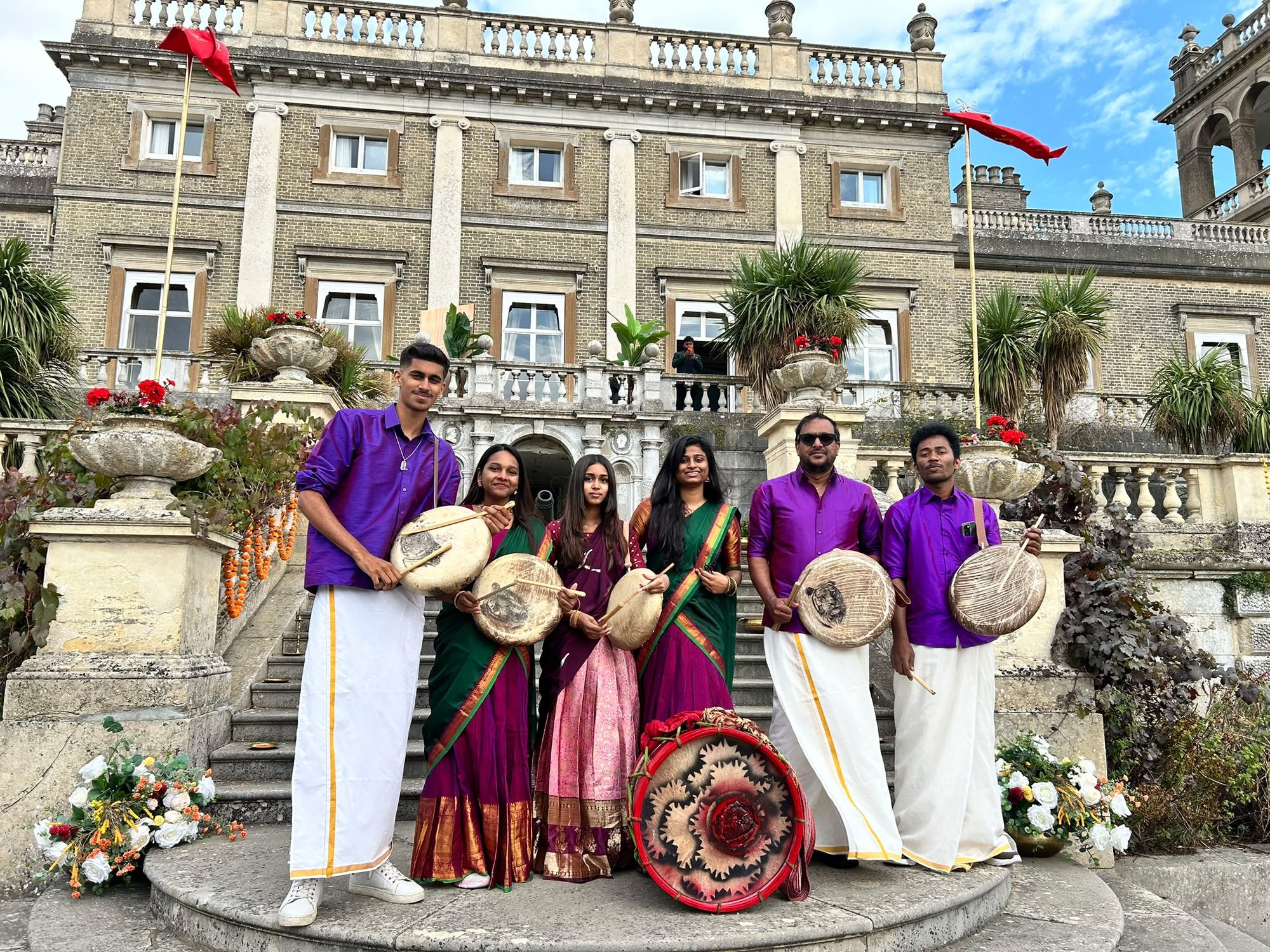 Seven Tamil Arts UK performers in purple kurtas and green and magenta sarees, holding parai drums on the steps of a stately home hung with red flags and marigold garlands