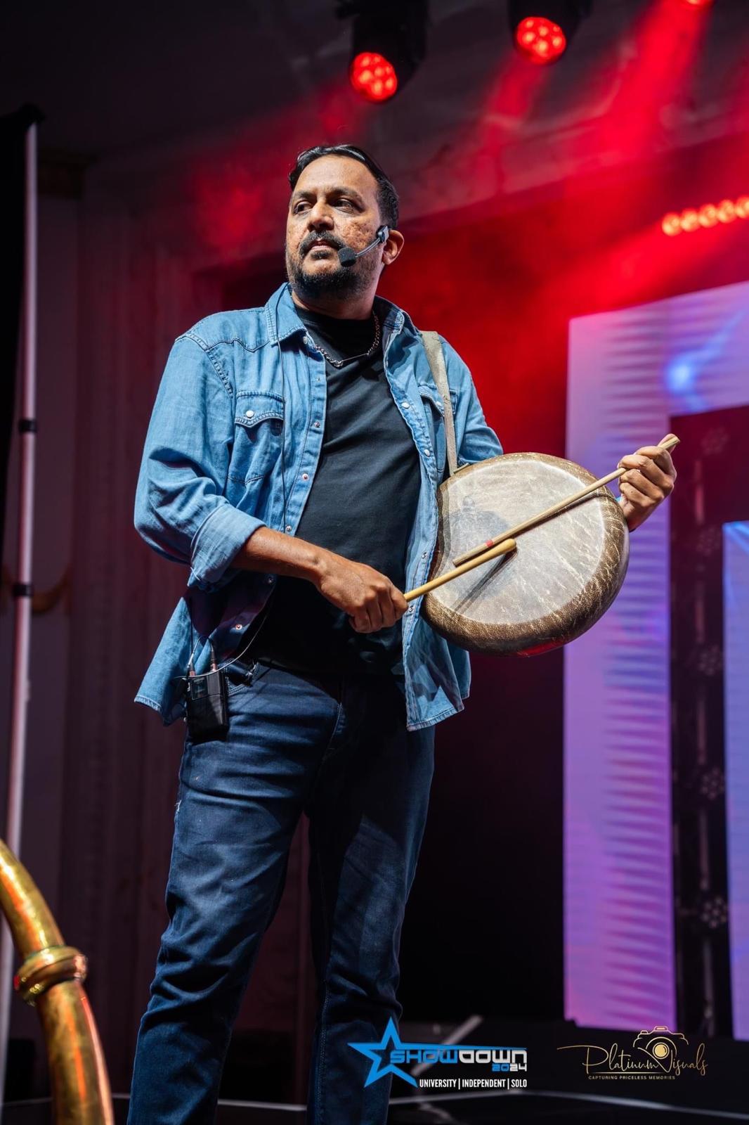 Karthik Kamalakannan playing the Parai on stage under red and blue lights, wearing a headset microphone