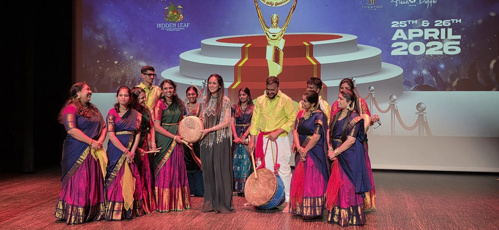 Tamil Arts UK performers on stage in silk saris and veshtis, holding parai drums and sticks, in front of an awards backdrop